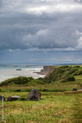 Panoramic view of the Normandy coast near Arromanches-les-Bains. Historic WWII Mulberry harbour remnants in the sea, green grassy cliffs and a dramatic cloudy sky on a stormy day in France.