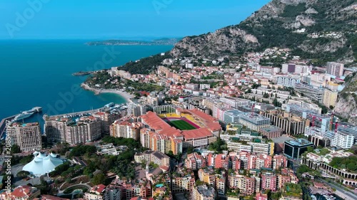 Aerial orbit over Stade Louis II football stadium in the Fontvieille district of Monaco, with the port, apartment buildings, coastal cliffs and the Mediterranean Sea on the French Riviera.