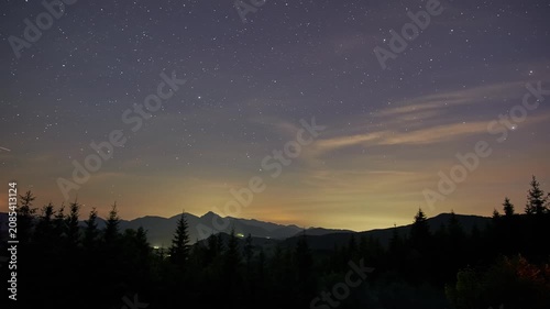Panoramic mountain nightscape showing shifting sky from warm horizon glow to cool blue with clouds and silhouetted evergreen forest