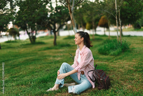 Fotografija Young woman sits on the grass in a park, wearing a pink plaid shirt and light jeans, enjoying a calm, reflective moment in nature, a soft breeze rustles nearby trees