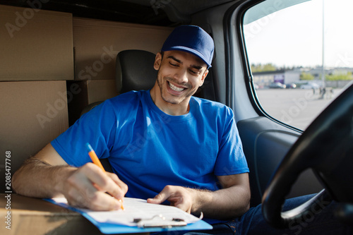Happy man courier smiling while writing on clipboard, planning route for addresses, sitting in van with cardboard boxes nearby