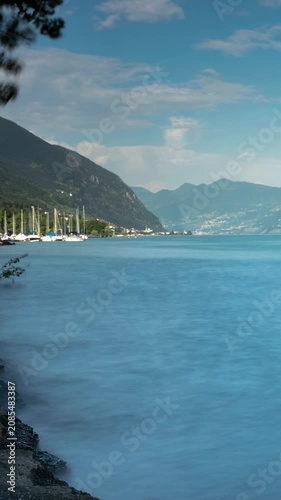 Time lapse of Lago d'Iseo lake with fluffy clouds moving across the blue sky over alpine mountains and sailboats moored at the rocky shoreline in Italy