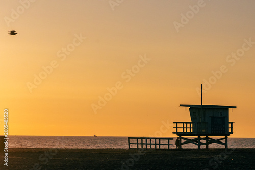Santa Monica Beach, California, USA - Silhouette of a lifeguard tower on Santa Monica Beach at sunset, with a seagull in flight and the Pacific Ocean on the horizon over water