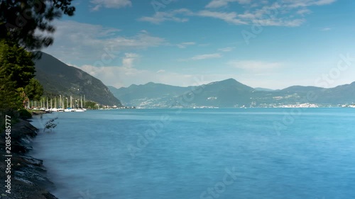 Time lapse of bright blue water of a mountain lake on a sunny day with rushing clouds moving over the distant green peaks and a harbor with boats