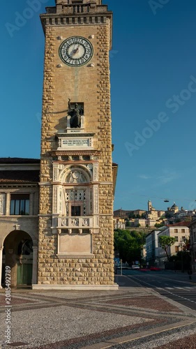 Timelapse of Torre dei Caduti clock tower with moving shadows and street traffic in Bergamo Italy against historic Citta Alta hill