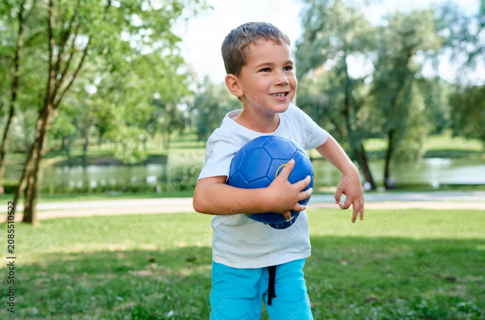 Obraz premium A cheerful little boy holds a blue ball while playing in a summer park.