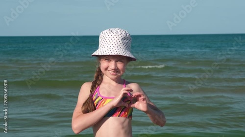 Happy girl in panama hat making heart gesture with hands on sea background. Concept of love for summer travel and holidays
