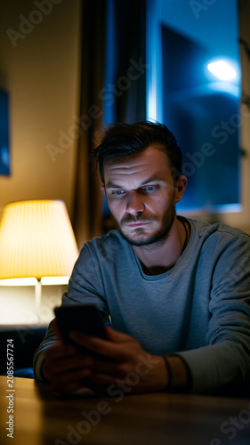 Concerned Man Looking at Smartphone in Cozy Living Room at Night