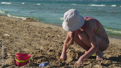 Girl in panama hat playing with sand near bucket on sea beach. Concept of creative children holiday activities and summer vacation