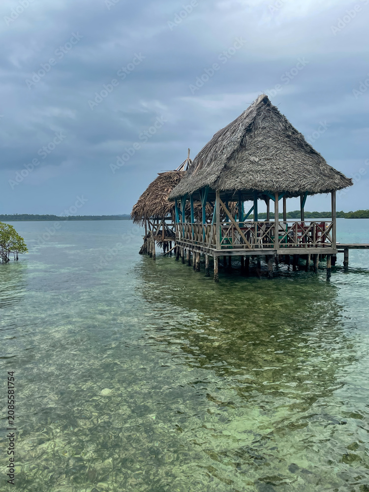 Fototapeta premium Thatched hut on wooden stilts over clear Caribbean waters near Bocas del Toro, Panama. Cloudy tropical sky