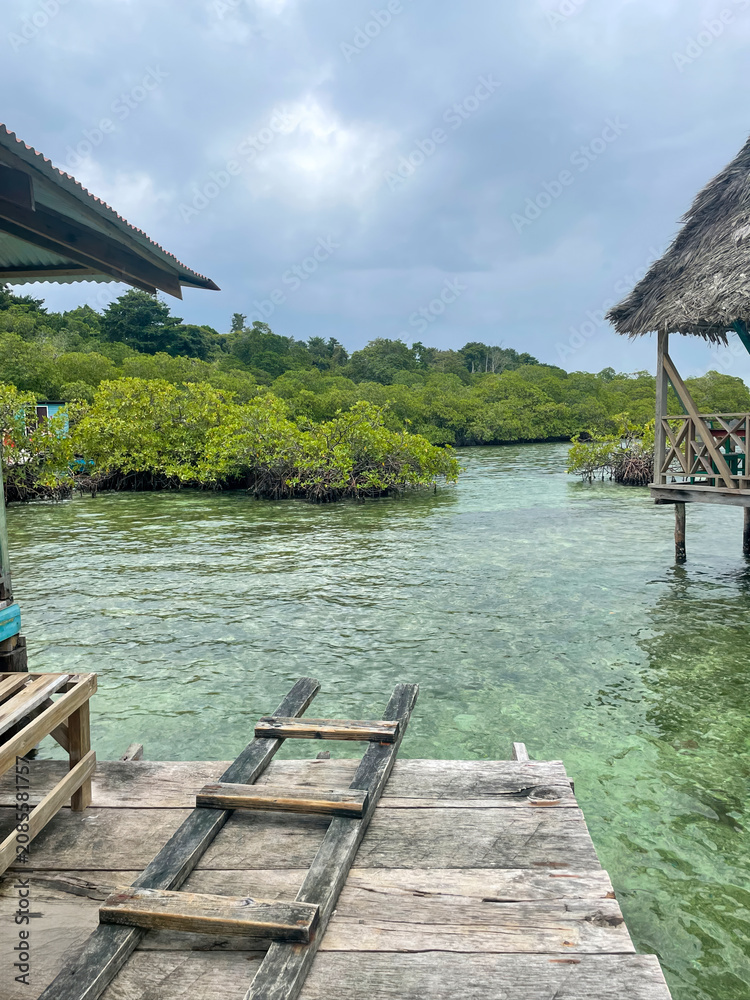 Fototapeta premium Wooden dock overlooking the calm mangrove lagoon within Bocas del Toro, Panama, under cloudy afternoon light featuring clear turquoise water and rustic huts creating a peaceful tropical mood