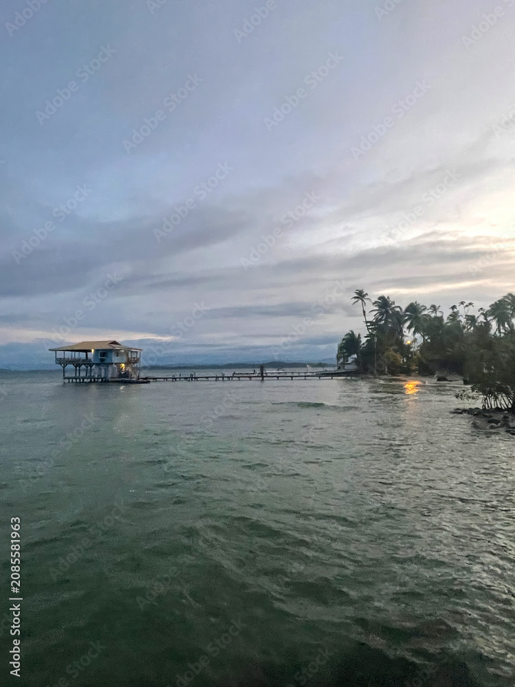 Fototapeta premium Palm trees on a sandy beach in Bocas del Toro, Panama at sunset. Wooden pier and stilt building, ocean waves, tropical sky.