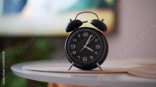 Black twin-bell alarm clock leaning slightly on round table near edge with soft warm indoor lighting, blurred background, centered clock face and vintage household style