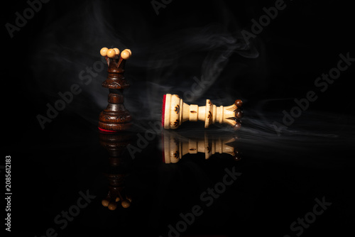 Fotografie Dark chess queen standing over fallen white queen in dramatic swirling smoke on black background