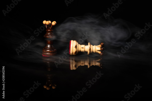 Fotografie Dark chess queen standing over fallen white queen in dramatic swirling smoke on black background