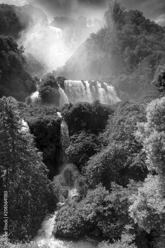 Vertical black and white view of Marmore Falls in Umbria, Italy. Multiple waterfalls cascade through dense forest while mist rises from the powerful water, creating a dramatic and timeless natural lan