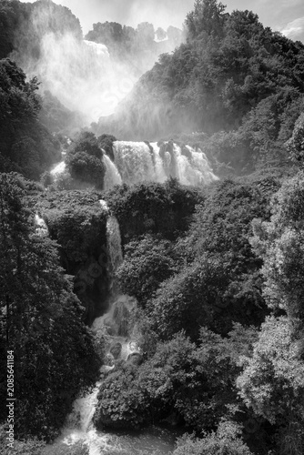 Vertical black and white view of Marmore Falls in Umbria, Italy. Multiple waterfalls cascade through dense forest while mist rises from the powerful water, creating a dramatic and timeless natural lan