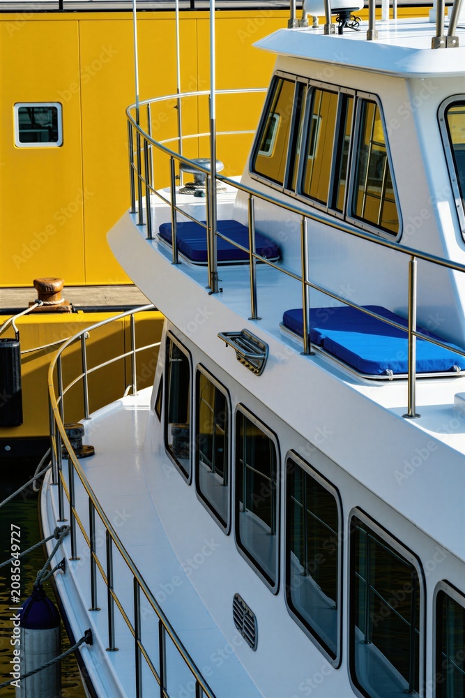 Fototapeta premium White boat docks next to bright yellow building. Vessel has blue cushions on deck. Metal railings line the ship sides. Windows reflect building and water.