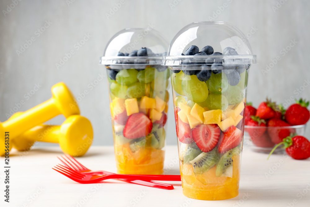 Fototapeta premium Tasty fruits in plastic cups, forks and dumbbells on light wooden table, closeup