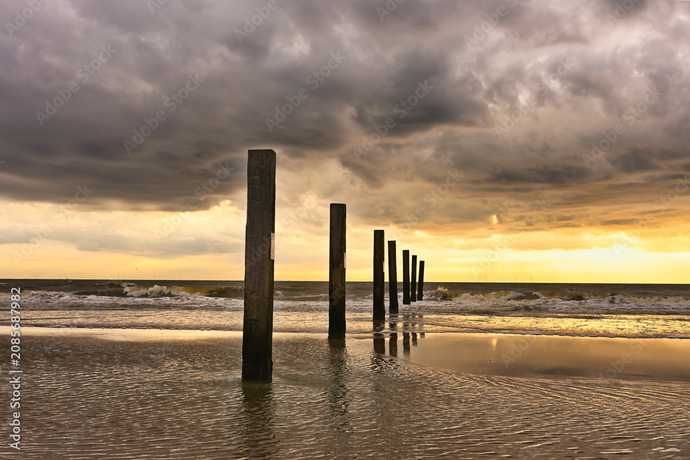 Obraz premium A warm sunset at Zandvoort with dramatic clouds, gentle waves, and wooden posts reflecting light on the wet sand for atmospheric designs.