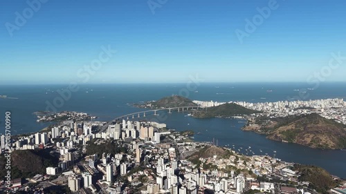 Aerial panoramic view of Vitória, Espírito Santo, Brazil, featuring Vitória Bay, islands, mountains, urban skyline, coastline, and clear blue sky captured by drone.