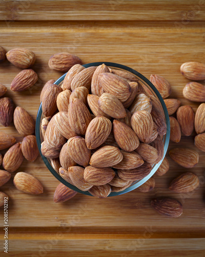 Natural almonds scattered around glass bowl on rustic wooden surface