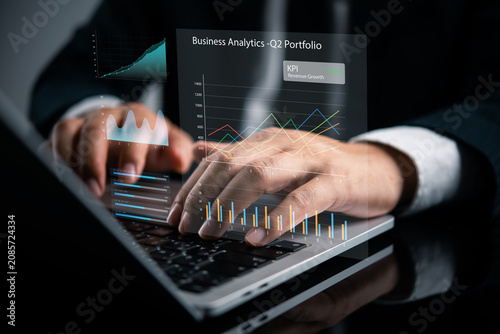 Businessman typing on laptop with virtual business analytics dashboard hologram displaying Q2 portfolio growth, revenue KPI, and strategic financial data for modern corporate success.