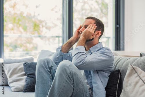 Bild auf Leinwand Young man sitting on a sofa at home, pressing his hands to his temples and showing signs of stress, headache and frustration in a bright, casual living room setting