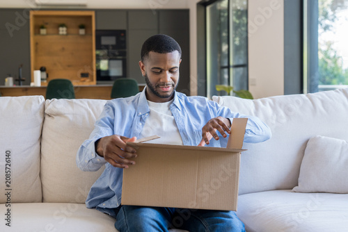 Εκτύπωση καμβά Young man sitting on a couch at home, opening a cardboard box received from an o