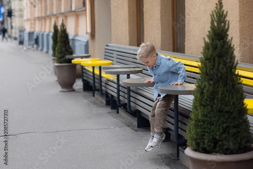 Foto Young boy in denim jacket playing on outdoor benches in Riga city, with potted p