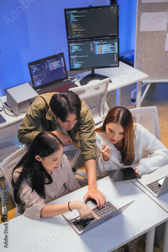 Three young Asian professionals - one man and two women - collaborate closely around a laptop, reviewing programming code together. Multiple monitors displaying code editors glow in the background 