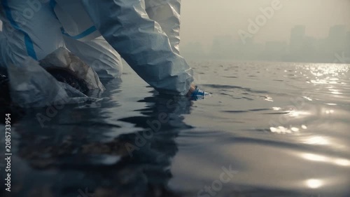 Close up shows worker removing waste from calm water near urban waterfront. Blue gloves and protective clothing emphasize environmental conservation and cleanup effort.