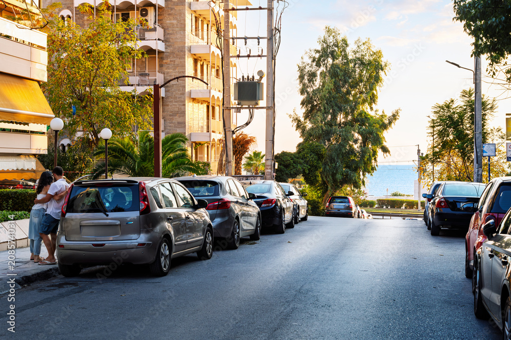 Fototapeta premium A young couple lean on a parked car on a seaside street with the Aegean Sea in the distance at dusk in the waterfront town of Palaio Faliro, Greece, on the Athenian Riviera.