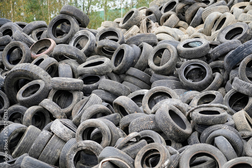 Hundreds of dark, worn vehicle tires are stacked haphazardly outdoors at a waste disposal site under daylight