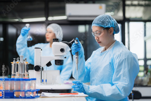 Person in blue lab attire adjusts microscope. Another individual holds test tube in background. Laboratory contains flasks and beakers. Modern setting with bright lighting