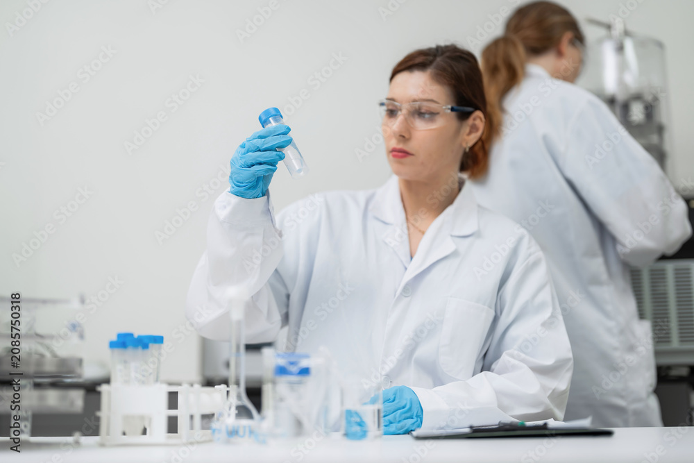 Fototapeta premium Woman in a white lab coat holds a test tube. She wears blue gloves and safety glasses. Lab equipment sits on the workbench. Another scientist works in the background
