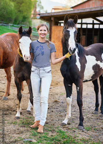 Young woman walking next to two American Paint Horses, blurred ranch with wooden boxes for animals in background