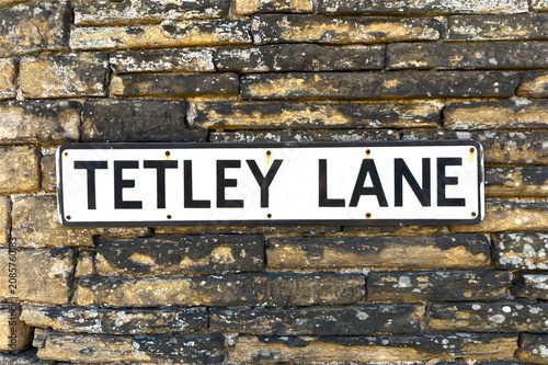 A vintage street sign reading 'Tetley Lane' rests against a textured stone wall, its weathered surface hinting at the charm and history of the old village of Shibden in Halifax, UK.