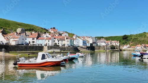 Panning shot across Staithes waterfront, harbour and fishing boats on the North Yorkshire coast, showcasing colourful cottages, sparkling water, and bright sunshine on a beautiful summer day.