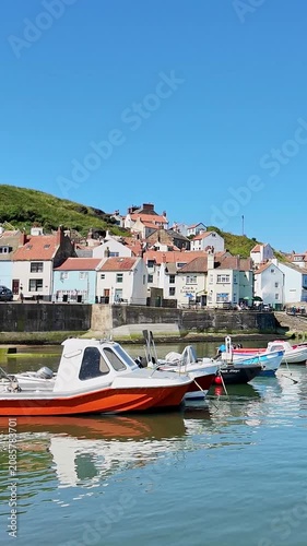 Panning shot across Staithes waterfront, harbour and fishing boats on the North Yorkshire coast, showcasing colourful cottages, sparkling water, and bright sunshine on a beautiful summer day.