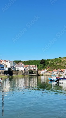 Vertical panning shot across Staithes waterfront, harbour and fishing boats on the North Yorkshire coast, revealing colourful cottages, sparkling water, and bright sunshine on a beautiful summer day.