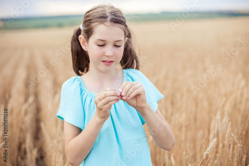 Young girl in light blue outfit stands in a wheat field, examining a piece of straw with a cloudy sky in the background