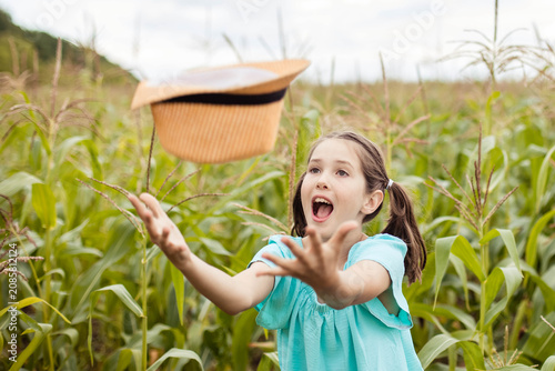 Young girl with brown hair wearing a blue shirt joyfully throws a straw hat into the air while standing in a lush green cornfield under a cloudy sky