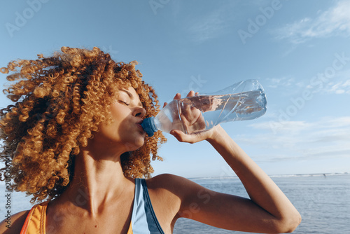Foto Woman with curly hair drinking water from a clear bottle by the sea under blue s