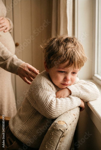 Sad little boy sitting by the window while mother stands nearby  
