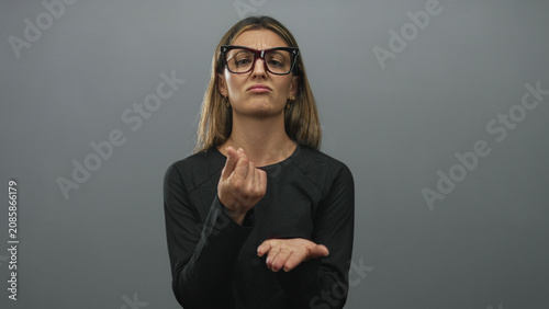 Fotografía Woman wearing glasses rubbing fingers and pointing to open palm in studio with grey backdrop; pleading help