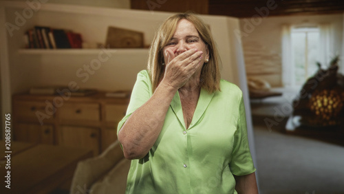 Photos Woman covering mouth with hand in a studio set resembling a living room with sofa and bookshelf; shock vulnerability