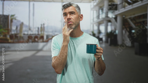 Fotografía Man holding teal mug presses cheek with wince and closed eyes for toothache inside a bright building concourse; discomfort morning routine