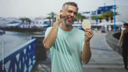 Tableau sur toile Man holding corn pastry and pressing finger to eye while sticking tongue out on street promenade; playful moment