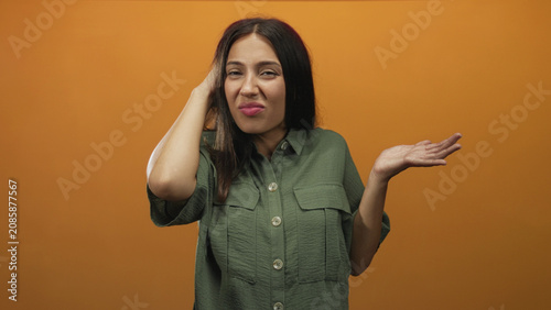 Foto Young brunette woman touching head with one hand and shrugging with the other in an orange studio; confusion